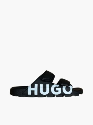כפכפי סליידס מעוצבים לגבר מבית HUGO בצבע שחור עם שתי רצועות סקוץ' ולוגו ענק לבן בצד – סטייל אורבני ונוחות מקסימלית.