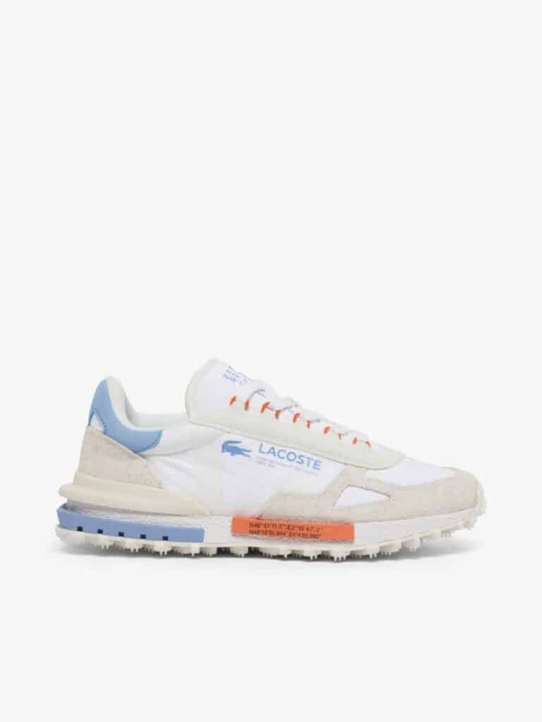 סניקרס לקוסט (Lacoste) בעיצוב ספורטיבי-מודרני, שילוב של ניילון לבן וזמש בז' עם נגיעות צבע בכתום ותכלת, כולל לוגו התנין וכיתוב קואורדינטות על הסוליה.