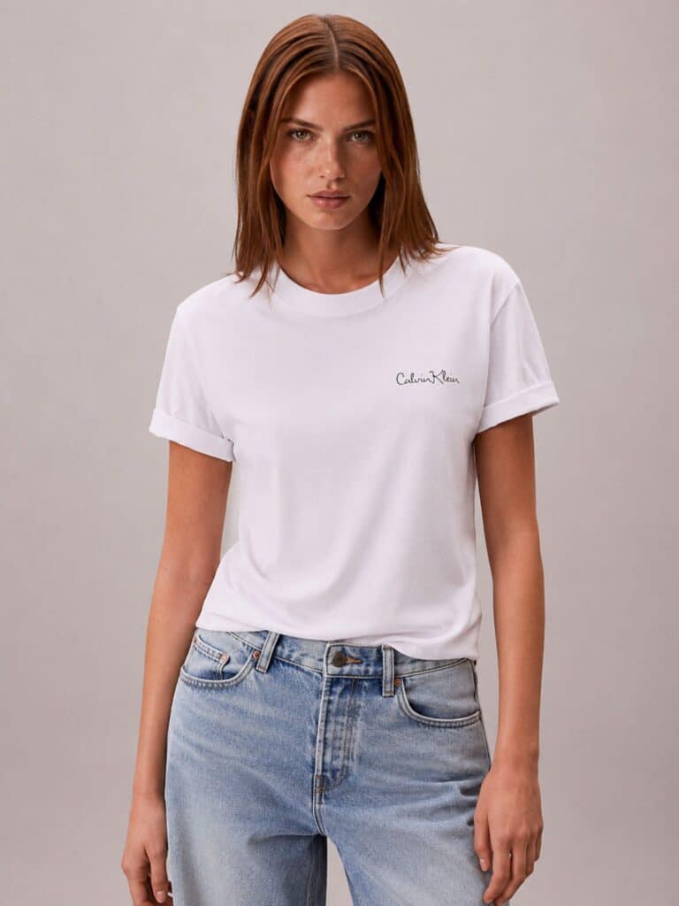 חולצת טי שירט לבנה לנשים מבית Calvin Klein, עשויה 100% כותנה בגזרת רגולר, עם לוגו המותג בכיתוב כתב-יד עדין בצד שמאל.